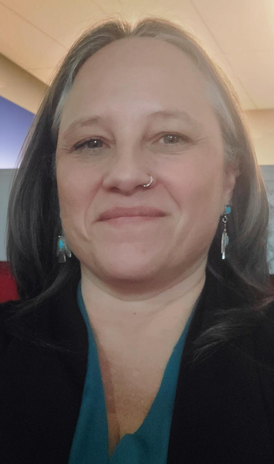 Angel Larkman; an Indigenous woman with long hair, turquoise earrings and black and teal clothing.